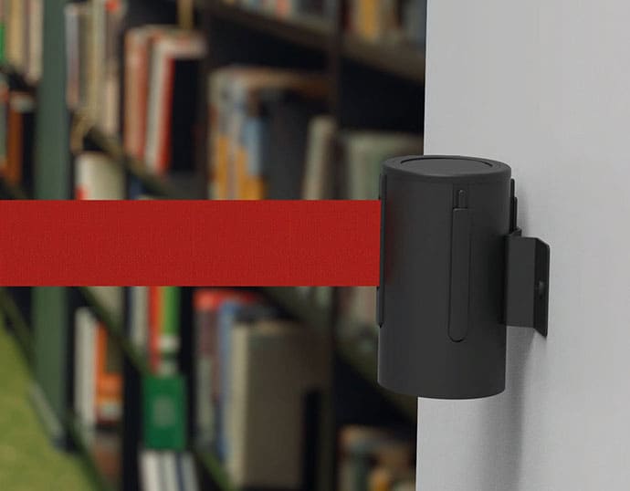 Las cintas separadores extensibles son muy habituales en bibliotecas, museos, centros comerciales, servicios de atención ciudadana, etc. Soporte mural con cinta separadora roja enganchado en pared de biblioteca