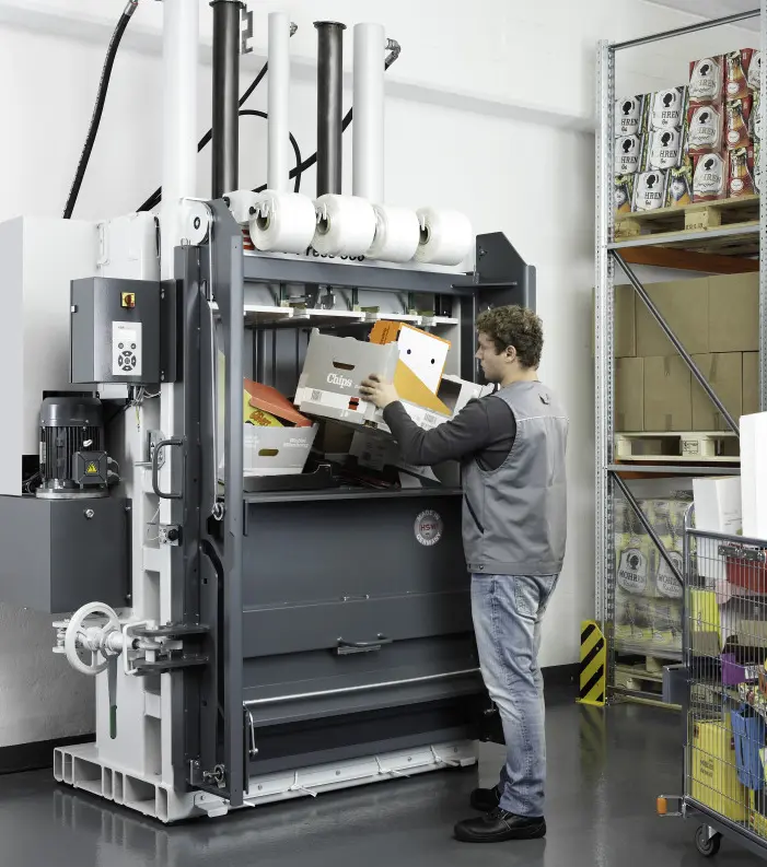 Trabajador en almacén industrial llenando de cartón una prensa compactadora hidráulica para reducir su volumen