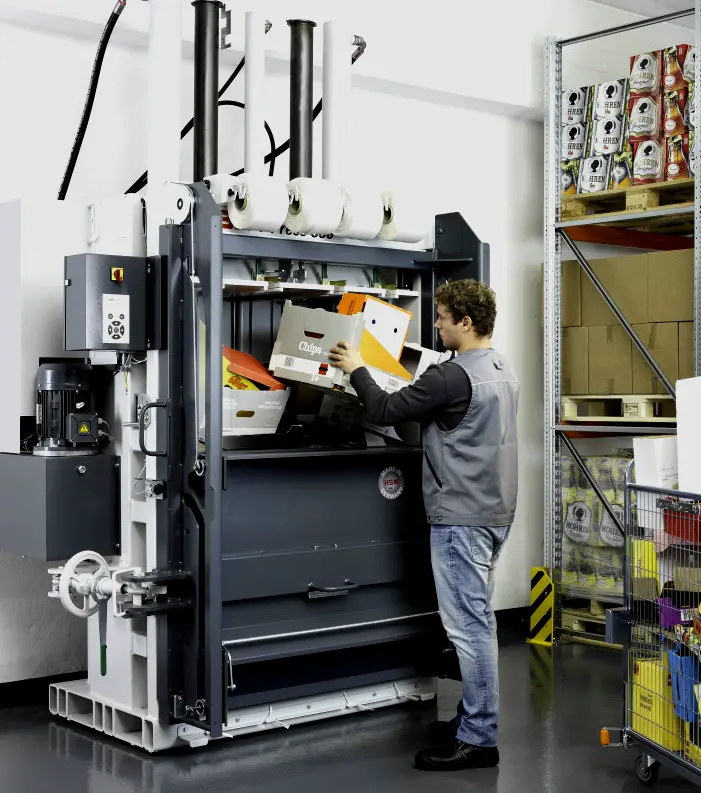 Trabajador en almacén industrial llenando de cartón una prensa compactadora hidráulica para reducir su volumen