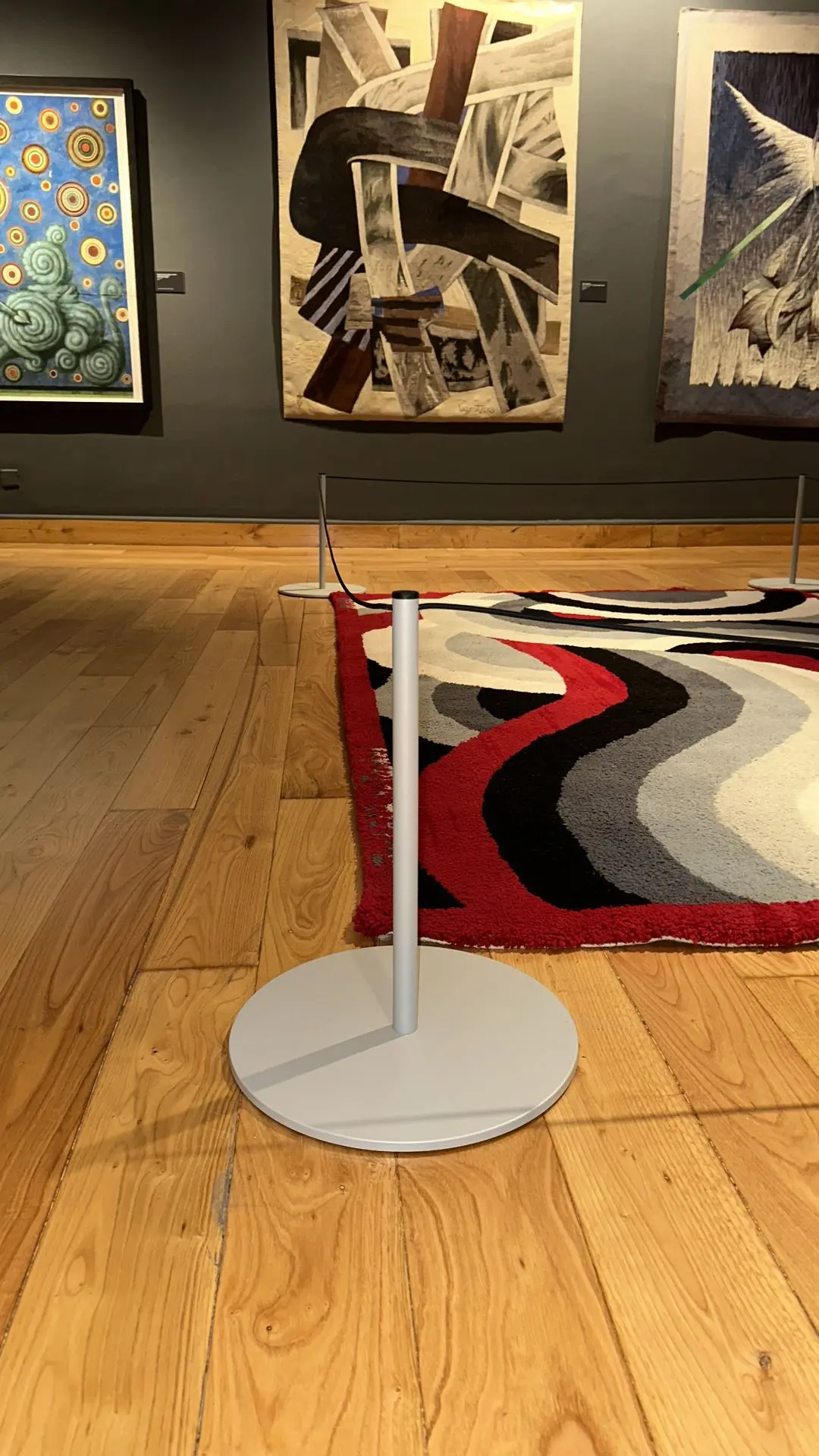 Detalle del poste Gallery Mini gris RAL 7035 con base de acero de 1 cm sobre parquet junto a una alfombra