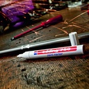 Detalle del Edding 750 escribiendo con tinta opaca sobre superficie metálica gris en entorno de trabajo industrial o técnico