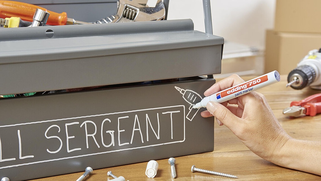 Marcador Edding 750 rotulando una caja de herramientas metálica con escritura opaca y trazo uniforme sobre superficie oscura