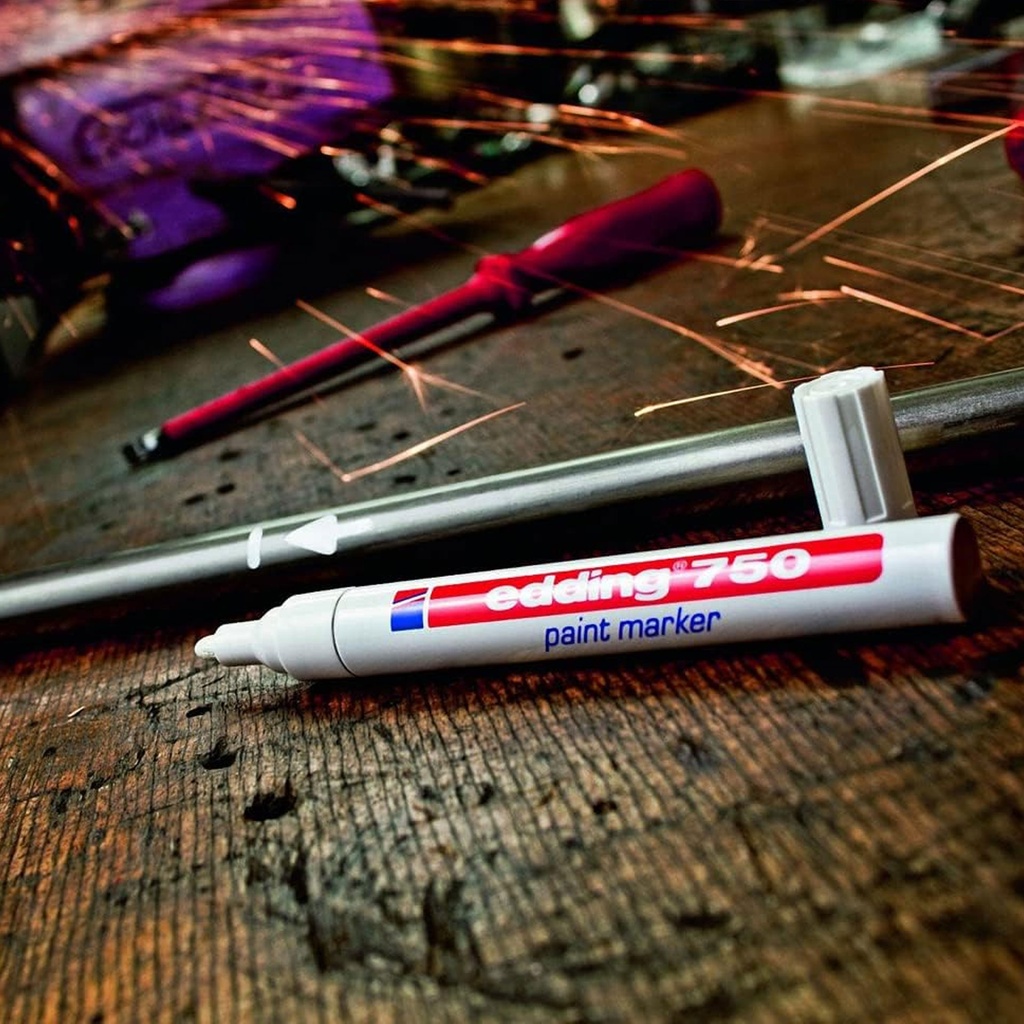Detalle del Edding 750 escribiendo con tinta opaca sobre superficie metálica gris en entorno de trabajo industrial o técnico